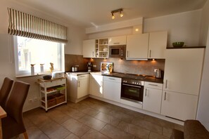 Eine moderne Küche mit weißen Schränken, einer Mikrowelle, einem Backofen und einer Kaffeemaschine. Ein Fenster mit Jalousien und eine kleine Ablage mit Dekorationselementen.