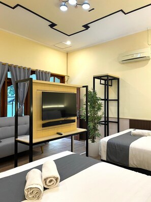 Habitación de hotel con televisor sobre un soporte de madera, sofá, cama con toallas, planta en maceta y unidad de aire acondicionado.