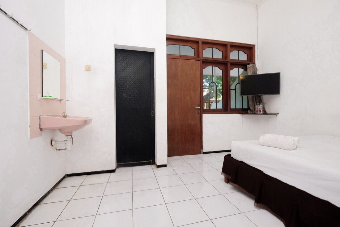 Una habitación pequeña y limpia con un lavamanos blanco, una puerta negra, una cama con una colcha blanca y un televisor montado en la pared.