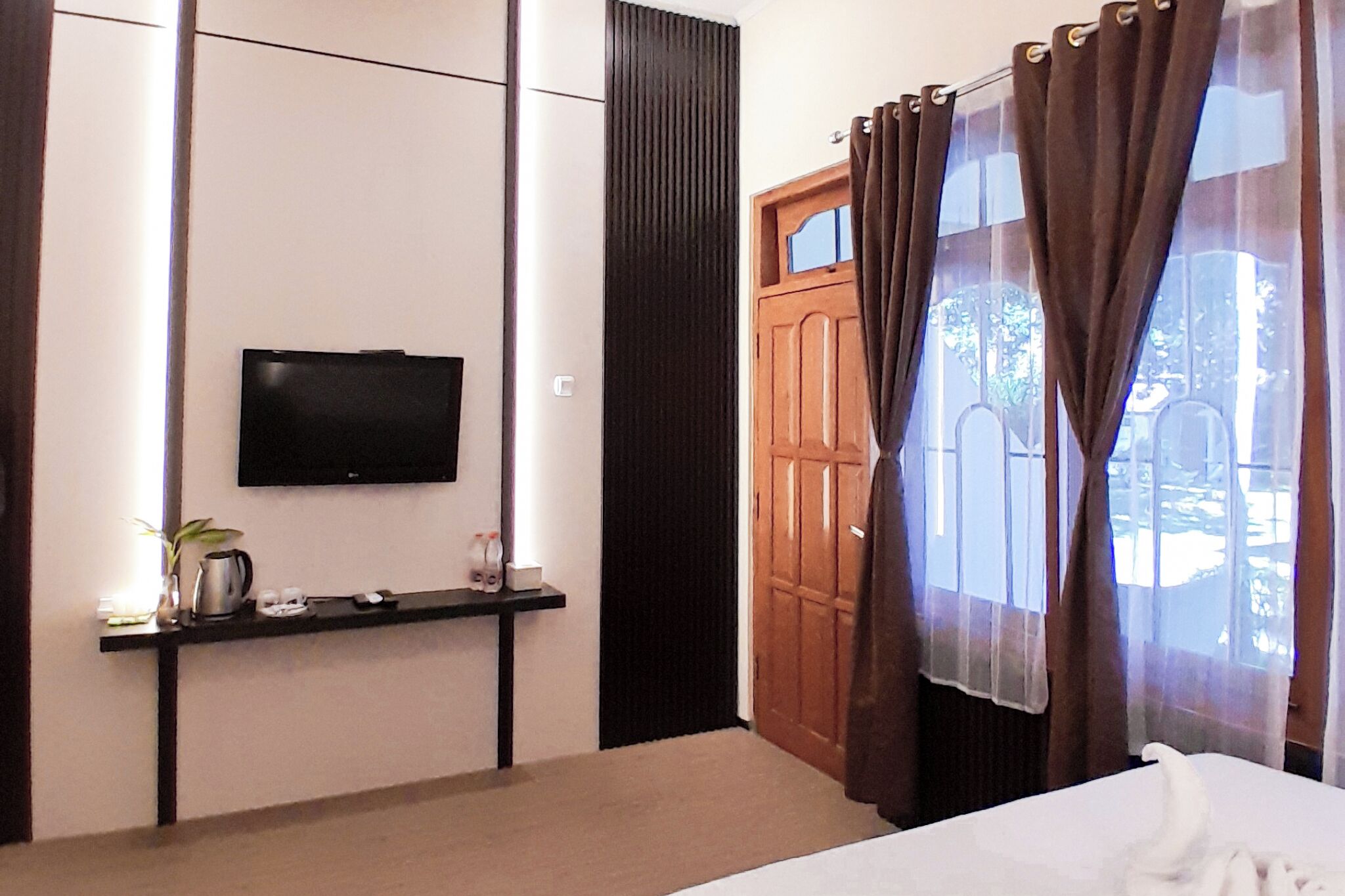 Habitación de hotel con cama, televisor montado en la pared, una mesita con planta y hervidor, y puerta de madera con cortinas.