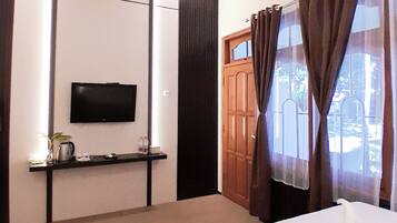 Habitación de hotel con cama, televisor montado en la pared, una mesita con planta y hervidor, y puerta de madera con cortinas.