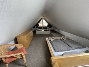 A small, triangular attic bedroom with a bunk bed, a wooden dresser, a wicker chair, and a window providing natural light.