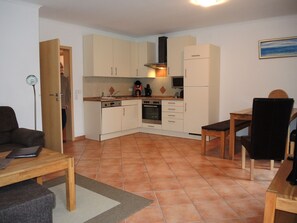 A compact kitchen with white cabinets, a stove, and a refrigerator. There is a dining table with chairs and a small rug on the floor. - Suite Anna with terrace 25 u (SA) - Usedom Suites 25***, beach 100 m (Karlshagen)