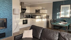 Eine moderne Küche mit weißen Schränken, einer Mikrowelle und einer Kaffeemaschine. Ein Esstisch mit Stühlen und einer Glasplatte steht im Raum. Ein Ledersofa mit Kissen ist im Vordergrund zu sehen.