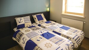 Una cama con ropa de cama azul marino y color beige con diseños náuticos, una ventana con persianas y una mesita de noche con una lámpara.