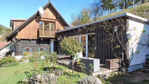 Una casa de madera de dos pisos con un balcón, una estructura pequeña adosada con puerta de vidrio y un jardín con diversas plantas.