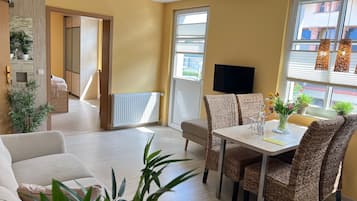 A modern living room with a sofa, dining table, wicker chairs, a TV, and potted plants.