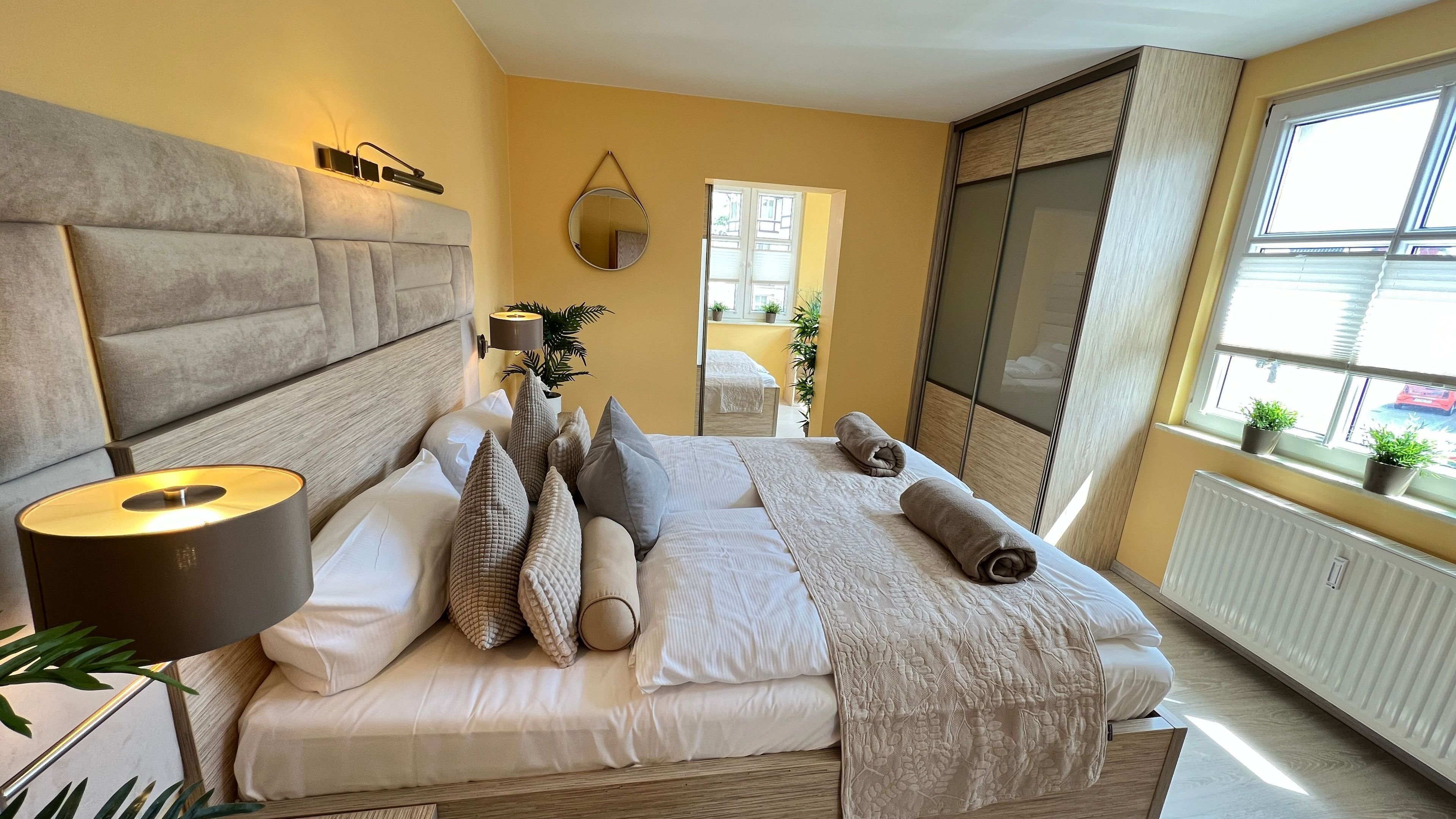 A bedroom with a large bed, a bedside lamp, a mirror, a radiator, and a window with plants.
