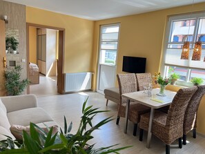 Una sala moderna con un sofá, mesa de comedor, sillas de mimbre, televisor y plantas en macetas.