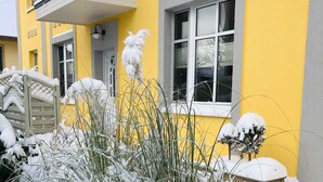 Um edifício amarelo com varanda, um portão de madeira e plantas cobertas de neve.