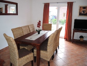 Comedor con mesa de madera, sillas de mimbre, televisor sobre un mueble y vista a través de un ventanal.