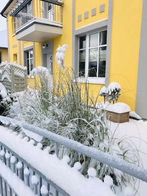 Een geel gebouw met een balkon, een houten hek en planten bedekt met sneeuw.