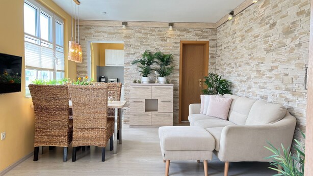 A modern living room with a sofa, dining table, wicker chairs, a kitchen area, and stone wall decor. - Apartment 1 ground floor - Villa Trautes Heim (Heringsdorf)