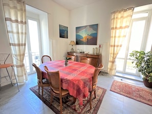 A dining area with a round table covered in a red tablecloth, surrounded by wooden chairs. There is a potted plant on the right side and a lamp on the table. - Charming Large T3 Bis in Port-Vendres, Ideal for Families (Port-Vendres)