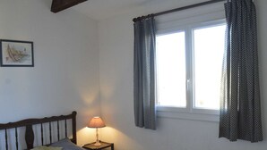 Ein Schlafzimmer mit einem Bett, einem Nachttisch mit Lampe, einem Fenster mit Vorhängen und einem gerahmten Bild an der Wand.