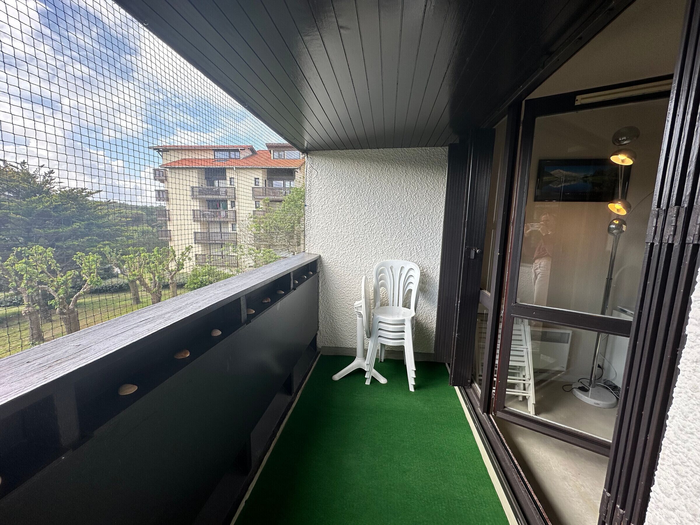 Un balcon doté d’un sol recouvert d’un gazon artificiel vert, d’une chaise en plastique blanche et offrant une vue sur un immeuble avec des balcons.