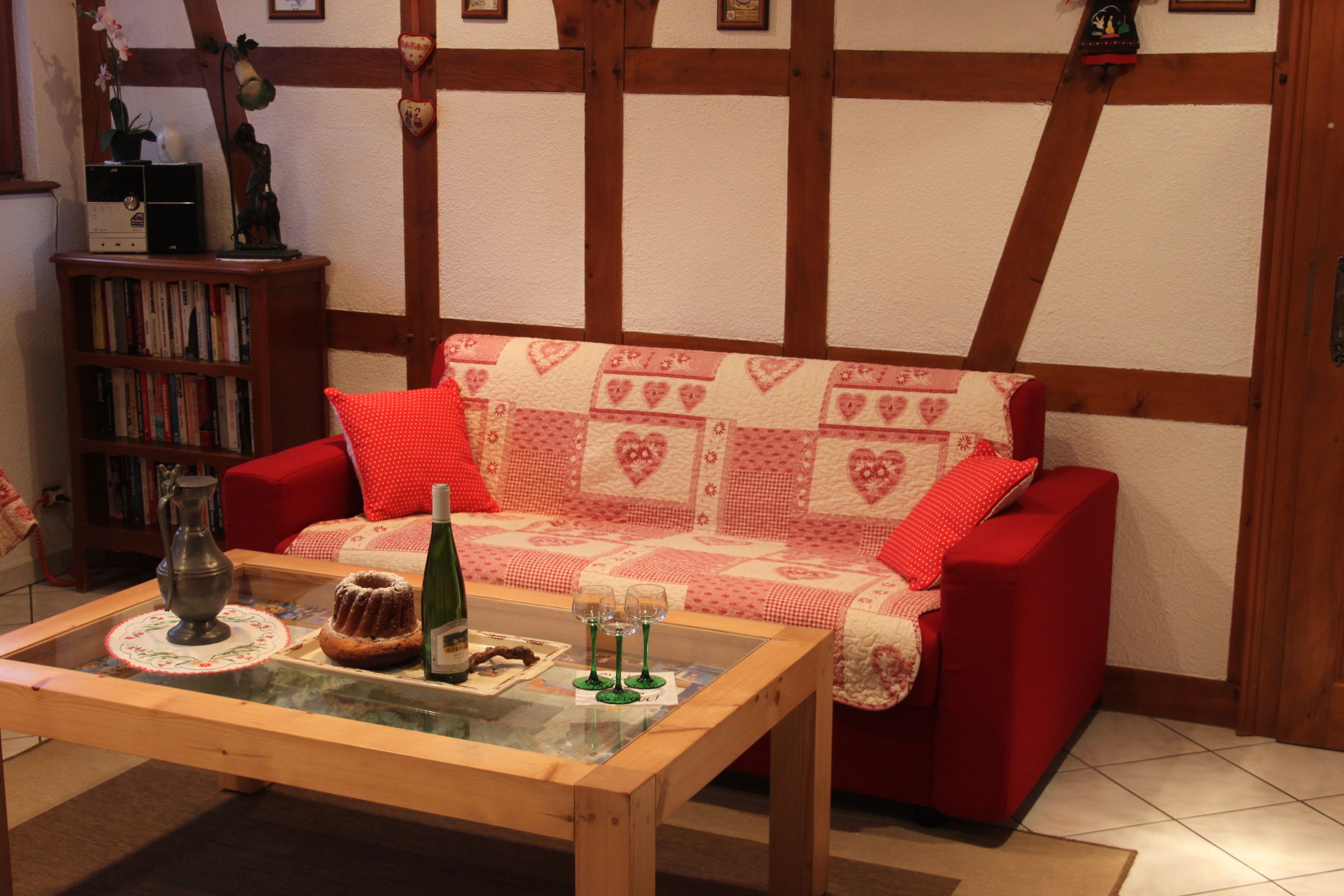 A cozy living room with a red sofa, a glass coffee table with a cake and wine glasses, and a bookshelf filled with books.