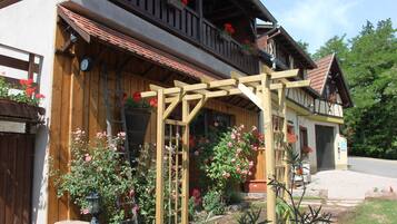 Une maison traditionnelle en bois, dotée d’un balcon, d’une pergola en bois et d’un jardin agrémenté de plantes à fleurs.