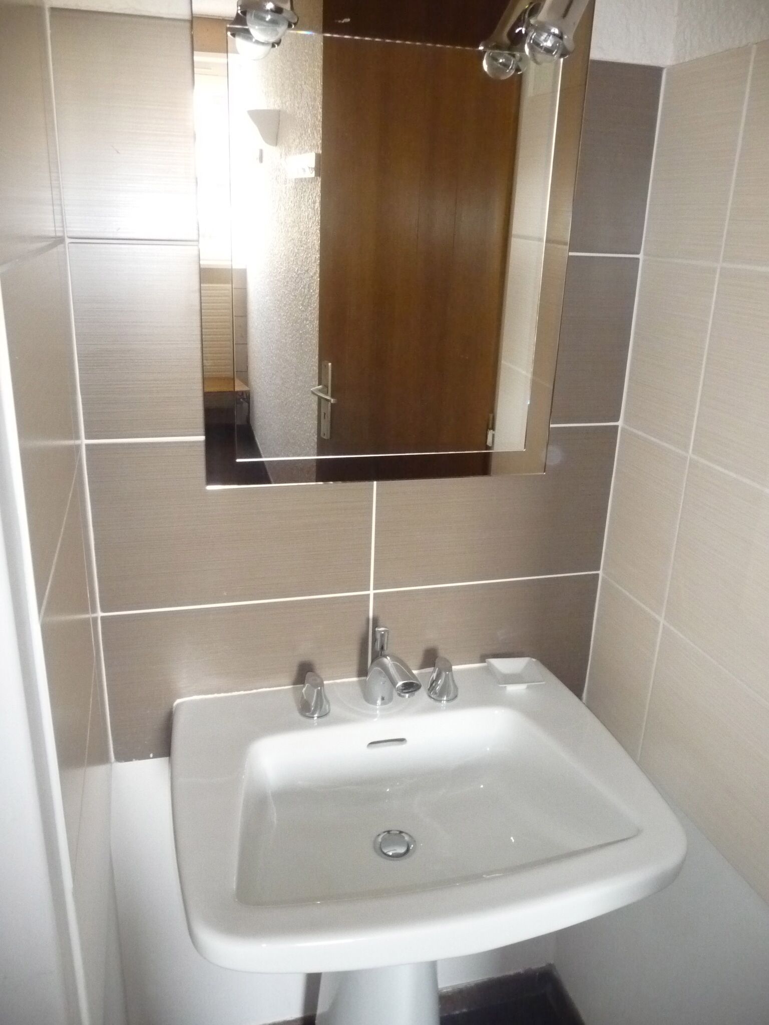 Ett badrum med ett vitt handfat, en spegel ovanför och beige kakel på väggarna.