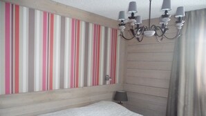 A hotel room with a striped wall, a wooden headboard, a bed with white bedding, a chandelier, and a window with curtains. - COURCHEVEL 1650, stand. apartment sleeps 8, 100 m gondola, covered parking. (Courchevel Moriond)