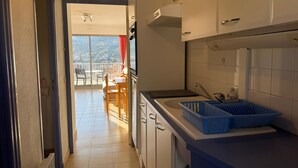 A narrow kitchen with white cabinets, a blue countertop, and a view of a balcony with a table and chairs. - Studio in Banyuls-sur-Mer, terrace close to beaches (Banyuls-sur-Mer)