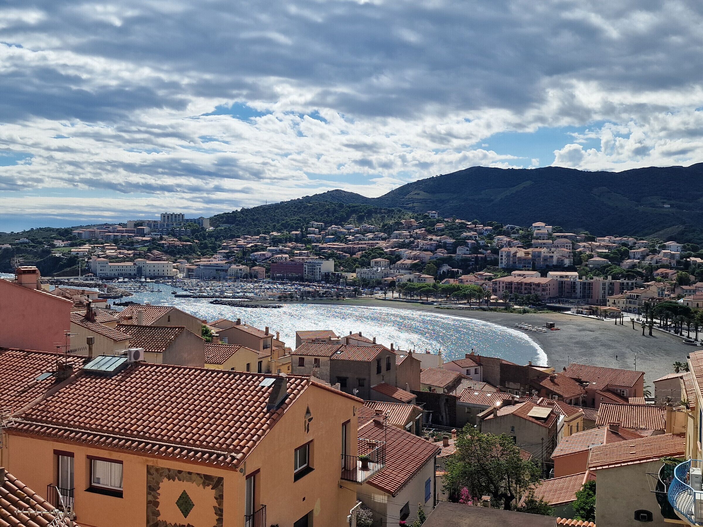 A coastal town with terracotta-roofed buildings, a marina, and a beach.