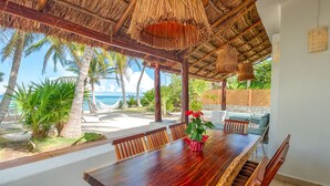 Een houten eettafel met stoelen, een rode bloevas en uitzicht op het strand met palmbomen en de oceaan.