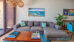 A modern living room with a grey sectional sofa, a wooden coffee table, and a painting of a beach scene. - Luxury beachfront ground level condo, perfect for a family, pool access, WiFi (AKUMAL)