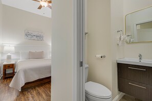 A bedroom with a bed, bedside table, and a nightstand. There is a bathroom with a toilet, sink, and mirror.