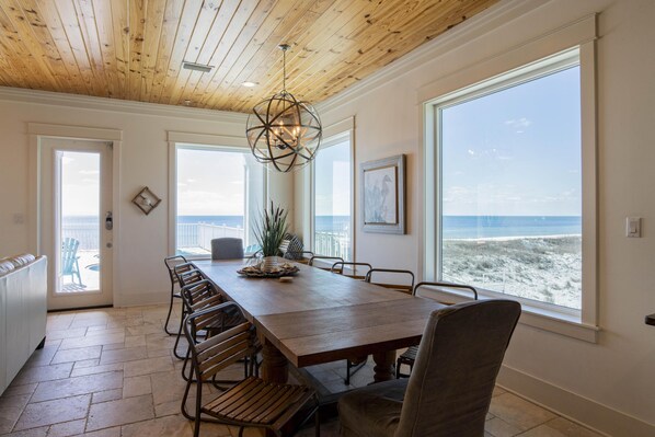 Un comedor con una mesa de madera, sillas y ventanas amplias que ofrecen vista a la playa.