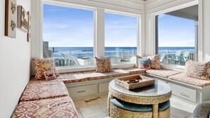 Una habitación soleada con un ventanal grande con vistas al mar, un banco tejido y cojines estampados.