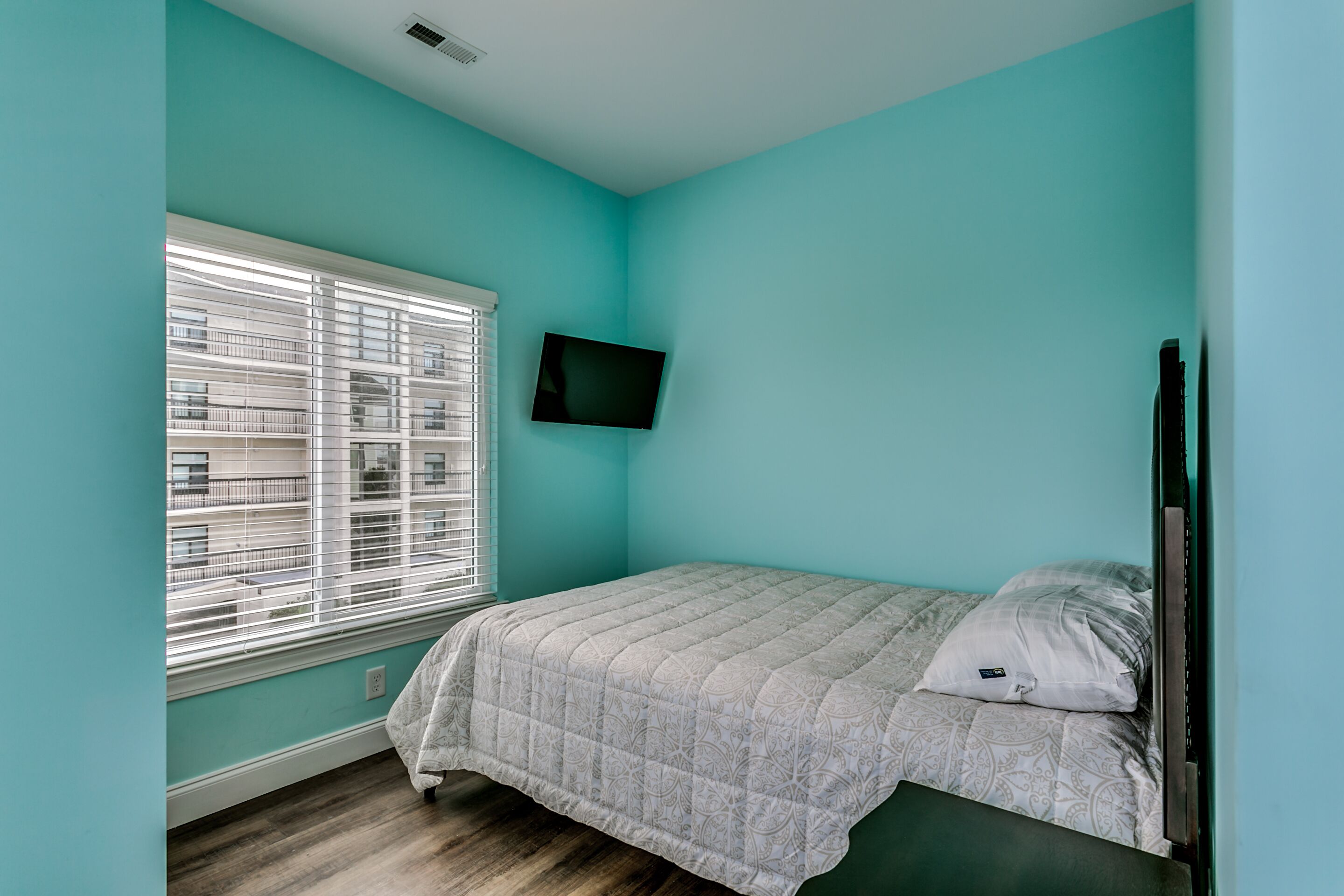 Een slaapkamer met turquoise wanden, een bed met een bedsprei met dessin, een wandtelevisie en een raam met jaloezieën.