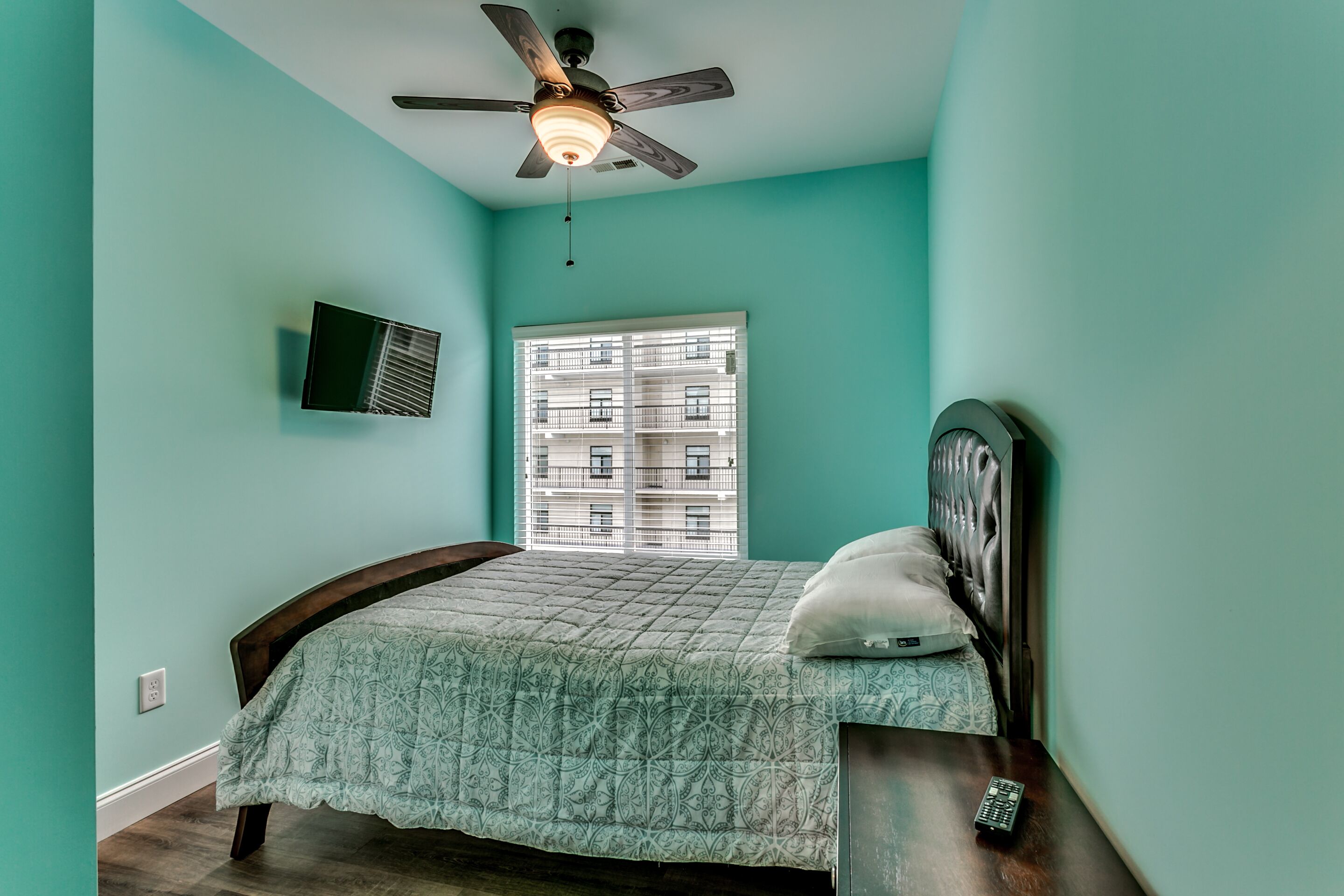 A bedroom with teal walls, a bed with a patterned bedspread, a ceiling fan, a window with blinds, and a remote control on the nightstand.