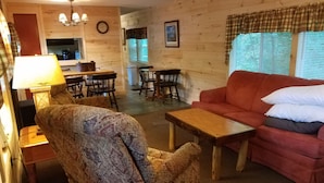 A cozy living room with a red sofa, patterned armchair, wooden coffee table, and a dining area with a table and chairs. - WATERFRONT and AFFORDABLE (Warrensburg)
