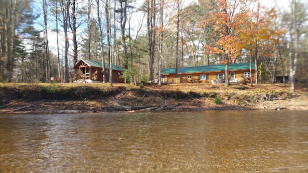 A cabin with a green roof and a wooden deck, surrounded by trees and a river. - WATERFRONT and AFFORDABLE (Warrensburg)