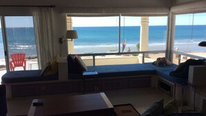 A living room with a large window overlooking a beach. There is a sofa, a small table, and a red chair. - Beach front in Rocky Point (Puerto Penasco)