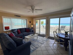 Un salon comprenant un canapé, une table basse et un coin repas avec des chaises. On y trouve un ventilateur de plafond, une décoration en forme d’étoile au mur et une vue sur la plage, visible à travers les portes-fenêtres coulissantes.