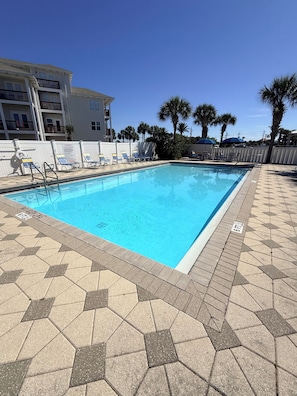 A swimming pool with hexagonal tiles, lounge chairs, palm trees, and a building with balconies.