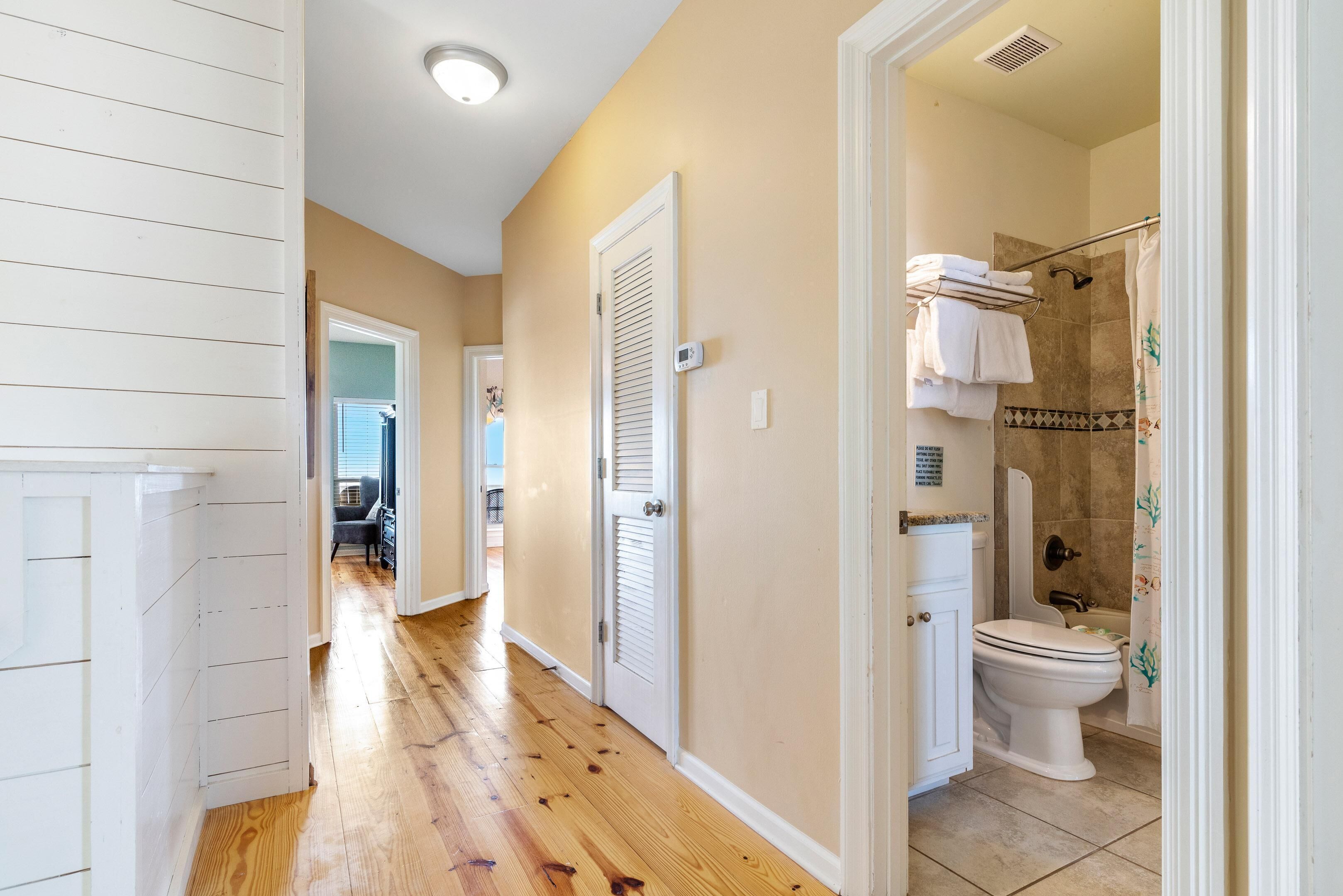 A hallway with a wooden floor, white walls, and a bathroom with a toilet, shower, and towel rack.