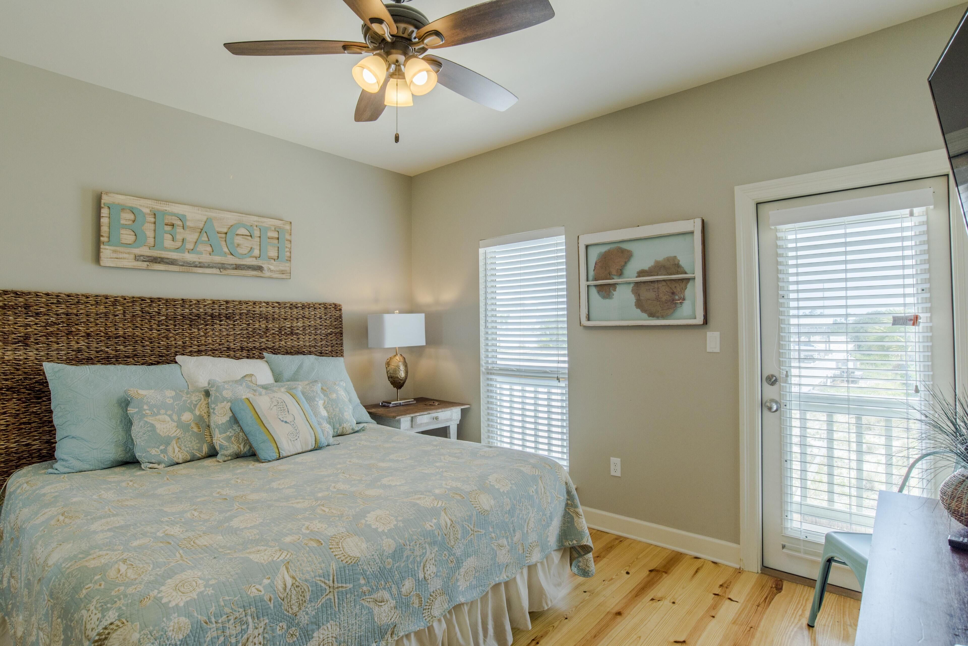 A bedroom with a bed, a ceiling fan, a bedside table with a lamp, a wall-mounted sign reading "BEACH", and a window with blinds.