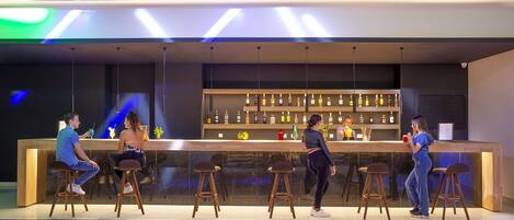 A modern bar with a long counter, multiple stools, and a variety of bottles on shelves behind the bar.