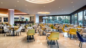 A spacious restaurant with multiple dining tables and chairs, large windows offering a view of the outdoors, and a modern ceiling design. - Club Marmara Doreta Beach Resort & Spa All Inclusive (Rhodes)