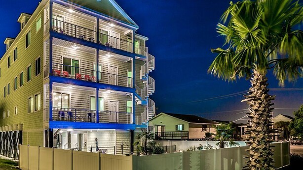 A multi-story hotel with balconies, a fenced yard, and a palm tree. - Private Retreat:master suite w Jacuzzi tub,game room,pool,outdoor entertainment! (North Myrtle Beach)