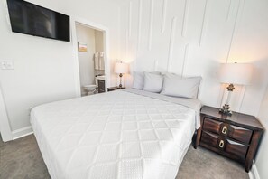 A bedroom with a large bed, two bedside tables with lamps, a TV mounted on the wall, and a door leading to a bathroom. - Private Retreat:master suite w Jacuzzi tub,game room,pool,outdoor entertainment! (North Myrtle Beach)