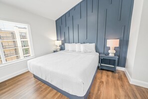 A bedroom with a large bed, a nightstand, two bedside lamps, a window with blinds, and a view of a building outside. - Private Retreat:master suite w Jacuzzi tub,game room,pool,outdoor entertainment! (North Myrtle Beach)