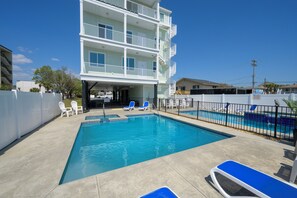 A swimming pool with lounge chairs and a multi-story building with balconies. - Private Retreat:master suite w Jacuzzi tub,game room,pool,outdoor entertainment! (North Myrtle Beach)