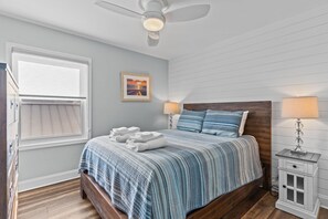 A bedroom with a bed, two bedside lamps, a ceiling fan, a window with blinds, and a framed picture on the wall. - Luxury Condo- Contemporary Oceanfront-Complete Remodel Keyless Checkin-RR Host (Carolina Beach)
