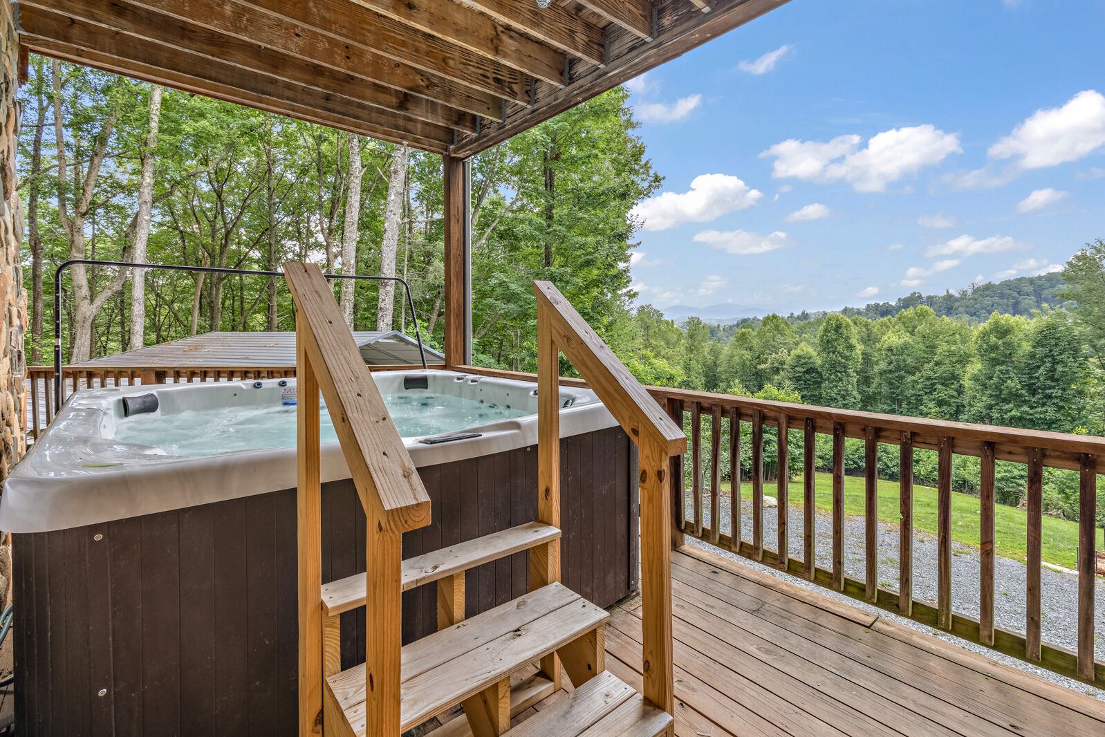 Une terrasse en bois dotée d’un jacuzzi et d’un escalier en bois menant à celui-ci, le tout entouré d’une balustrade en bois et offrant une vue panoramique sur les arbres et les collines.