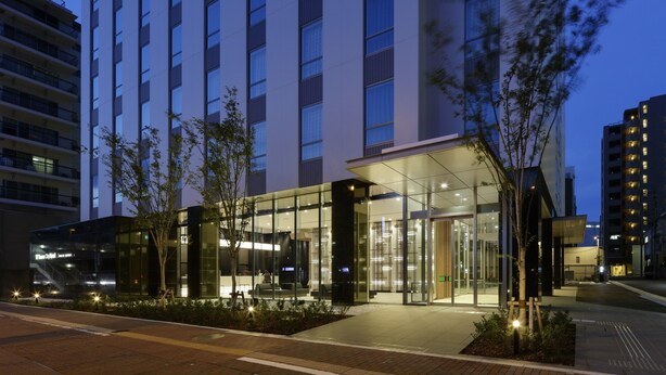 Una entrada de hotel moderna con puertas de vidrio, un área de espera cubierta y un jardín ajardinado.