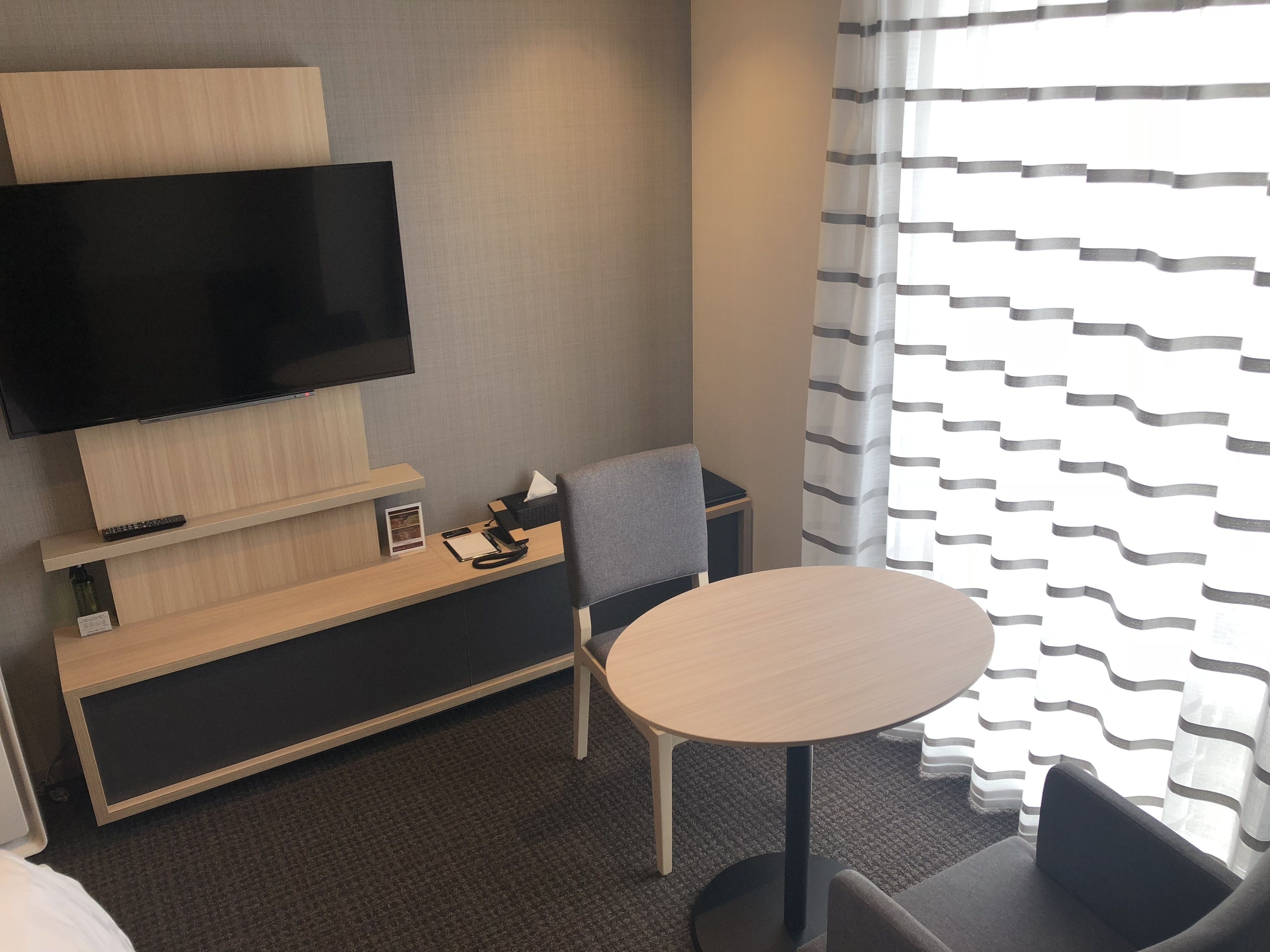 Habitación de hotel con televisor de pantalla plana montado en la pared, un escritorio pequeño con un teléfono y una libreta, una mesa redonda y un sillón gris.